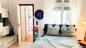A bedroom with a bed, a desk, a chair, a window with curtains, and a door leading to another room.