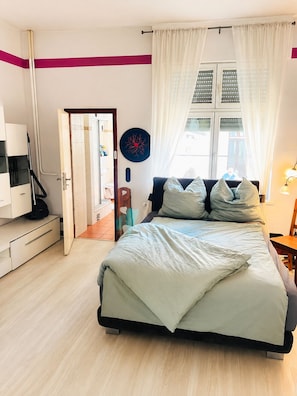 Ein Schlafzimmer mit einem Bett, einem Schreibtisch, einem Stuhl, einem Fenster mit Vorhängen und einer Tür zu einem anderen Raum.