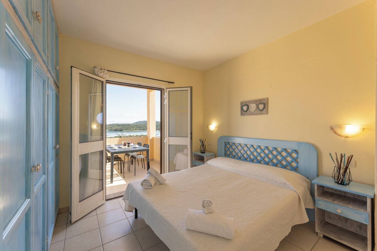 Une chambre à coucher avec un lit, une table de chevet, une table avec des chaises, et une vue sur la mer par une porte ouverte.