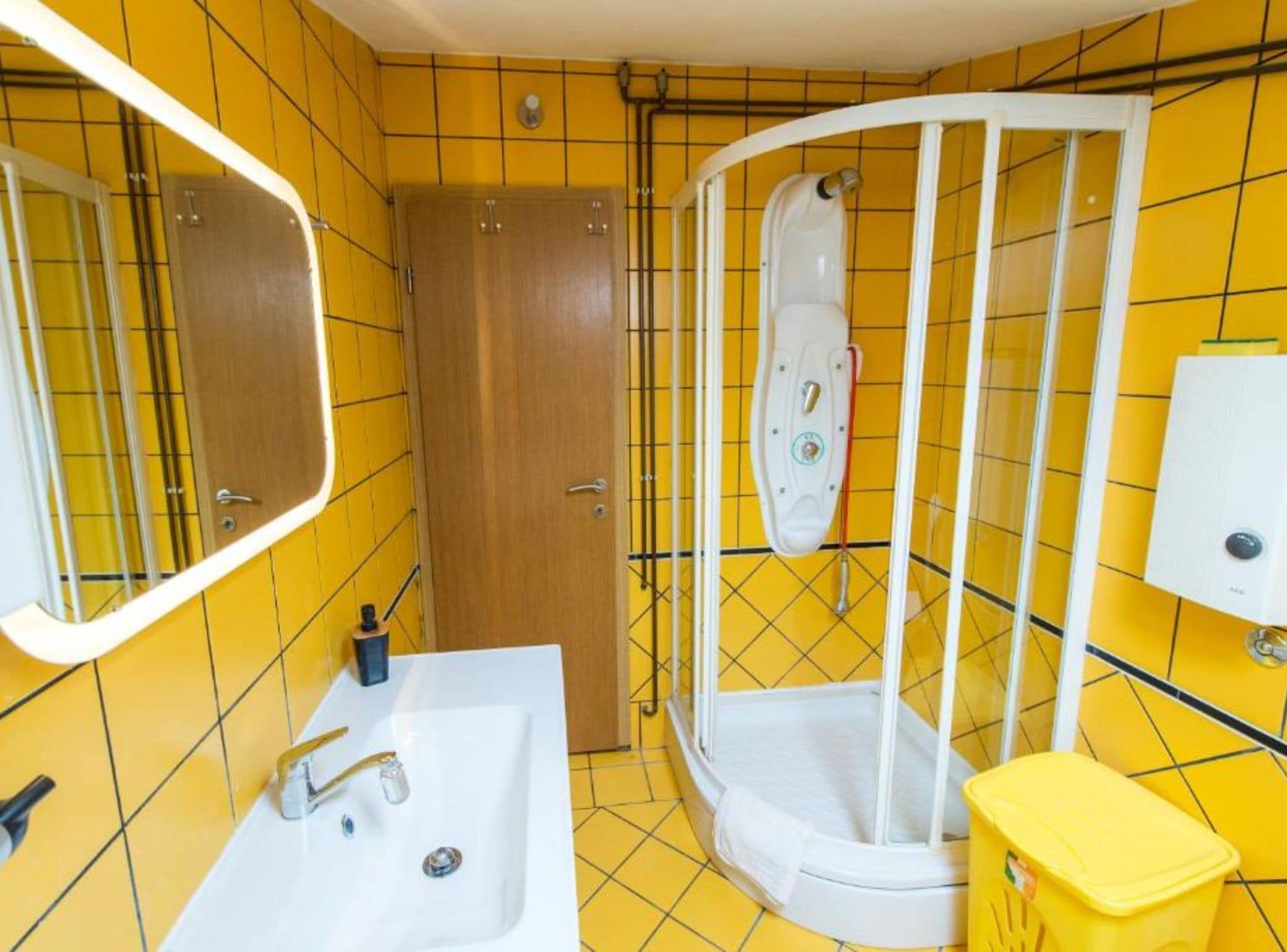 Een badkamer met geel betegelde wanden, een witte wastafel, een houten deur en een douchecabine.