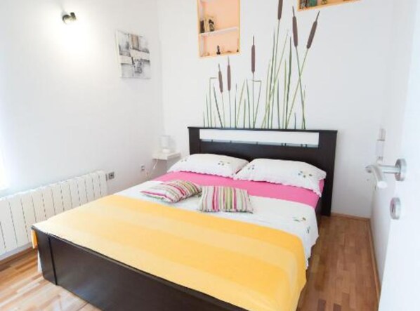 A bedroom with a bed, bedside tables, a radiator, and a wall-mounted shelf with decorative items. - Apartments Galić (Ploce)