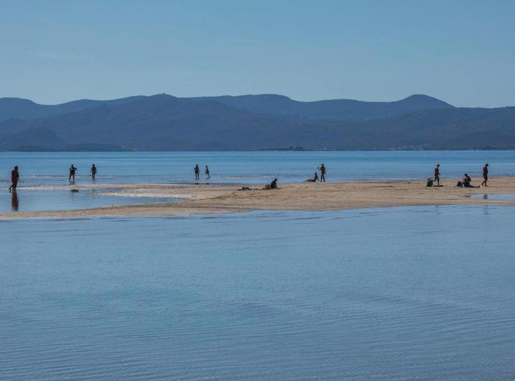 Una playa con gente caminando y sentada, un mar tranquilo y montañas al fondo.