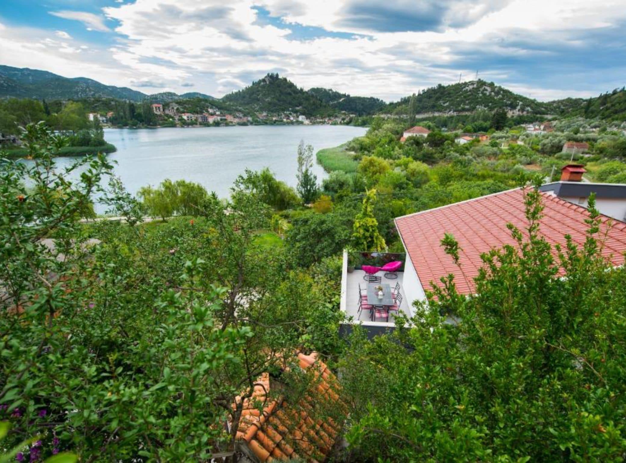 A view of a lake with a red-roofed building in the foreground, surrounded by greenery and hills in the background.