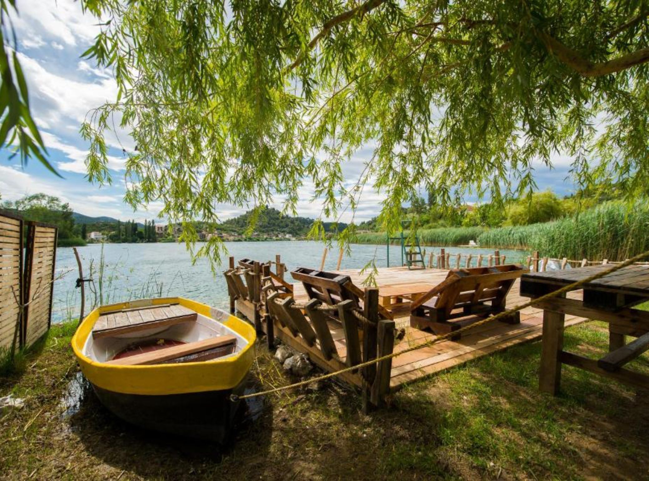 Un muelle de madera con un barco amarillo, bancos de madera y una mesa bajo un sauce junto a un lago.