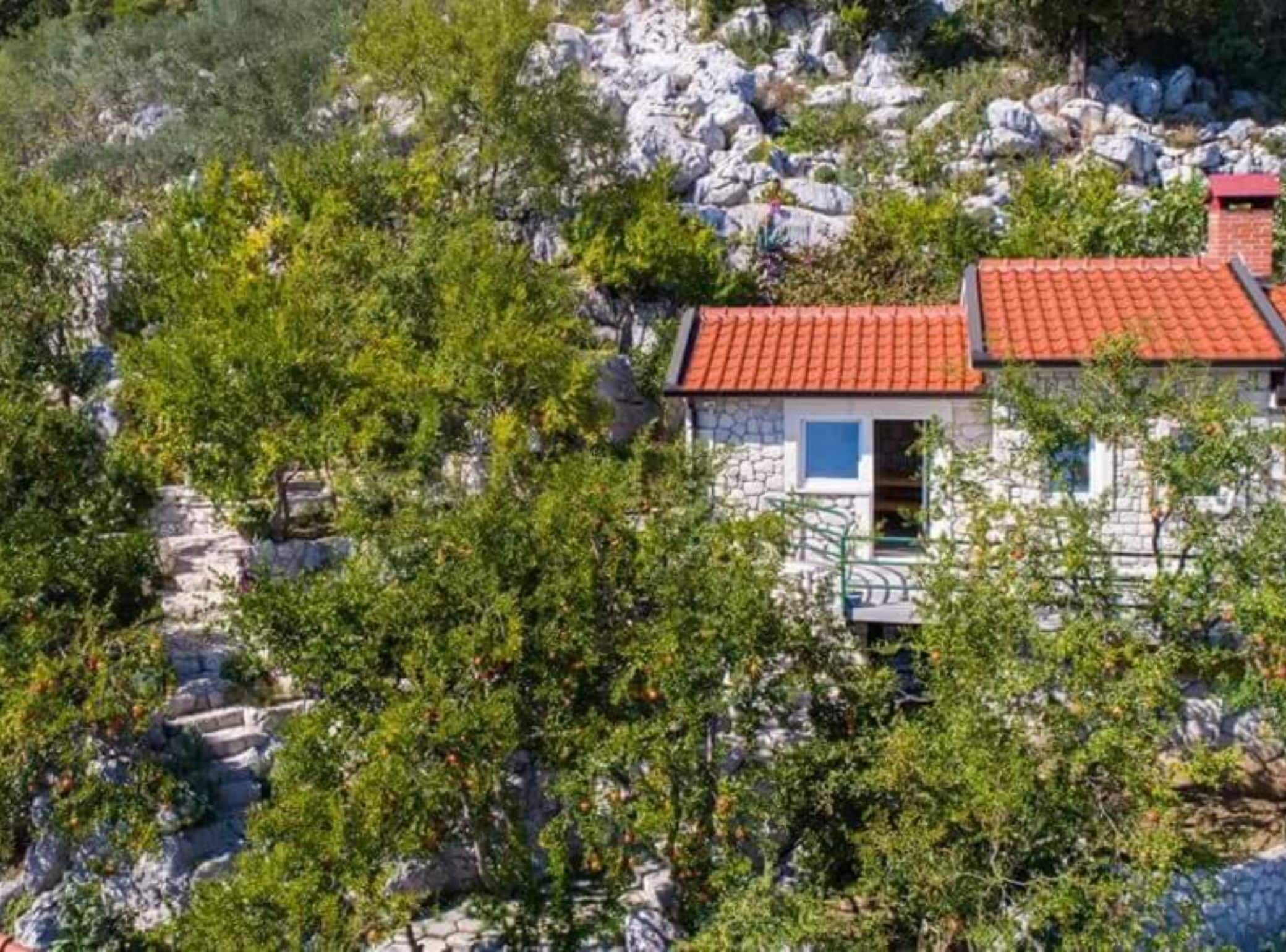 Een stenen huis met een rood pannendak, omringd door begroeiing en rotsen.