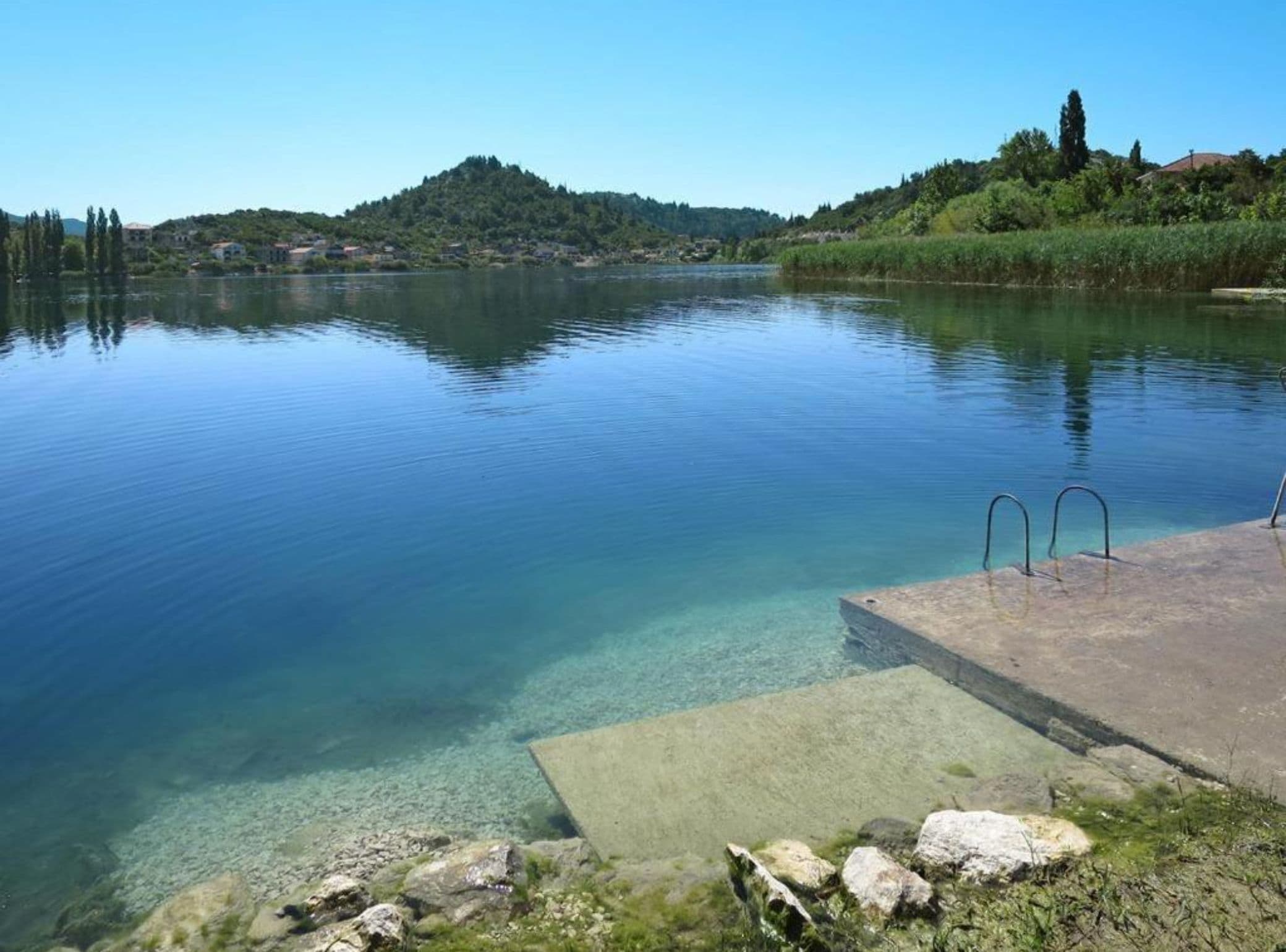 Un lago tranquilo con aguas cristalinas, un muelle de hormigón con barandillas metálicas y un paisaje montañoso al fondo.