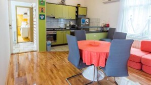 A compact kitchen with green cabinets, a dining area with a red table and blue chairs, and a red sofa. - Apartments Galić (Ploce)