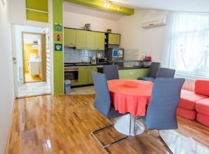 A compact kitchen with green cabinets, a dining area with a red table and blue chairs, and a red sofa. - Apartments Galić (Ploce)