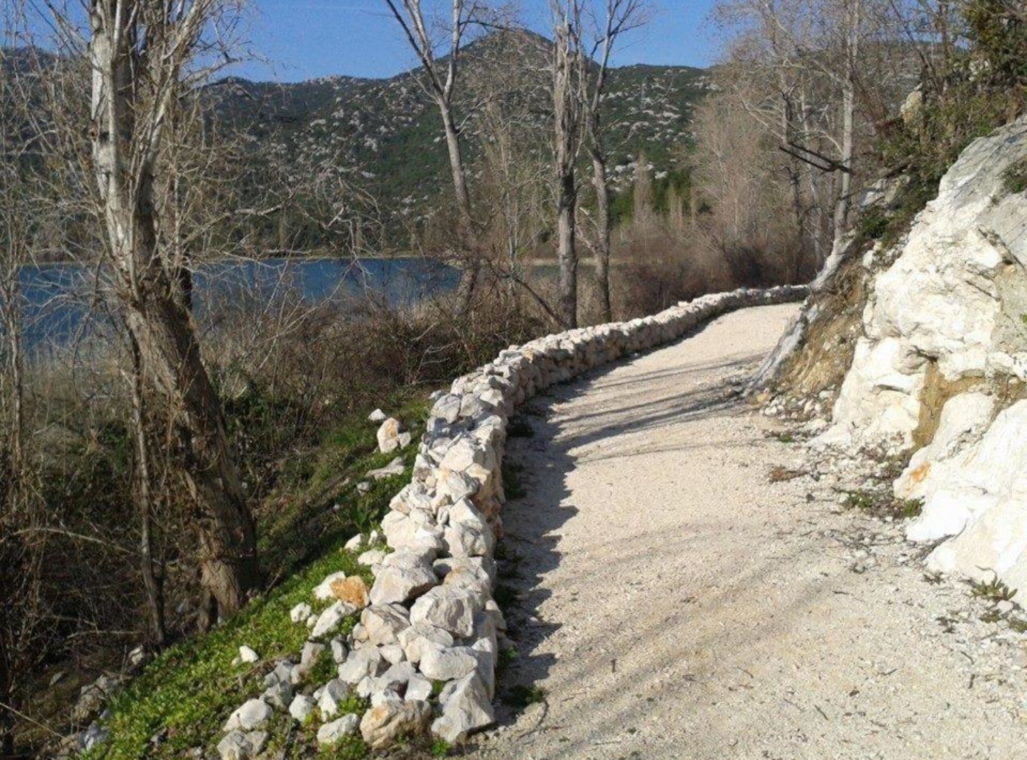 Un sendero de grava flanqueado por un muro de piedra, rodeado de árboles y una masa de agua al fondo.