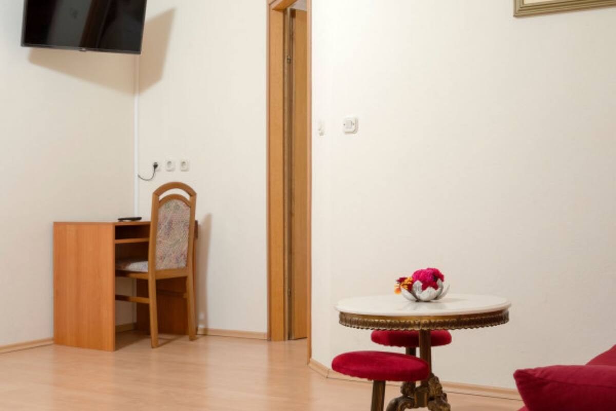 Una stanza con una televisione a schermo piatto montata a parete, una sedia di legno con cuscino decorato, un tavolino rotondo con cuscino rosso e un divano rosso.