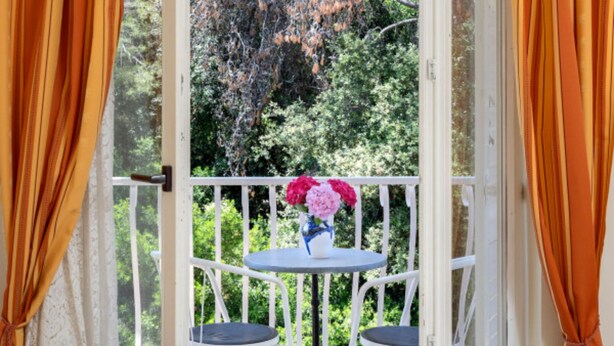 Ein offener Balkon mit weißen Stühlen und einem Tisch, eine Vase mit Blumen und Blick auf Bäume und Grün.