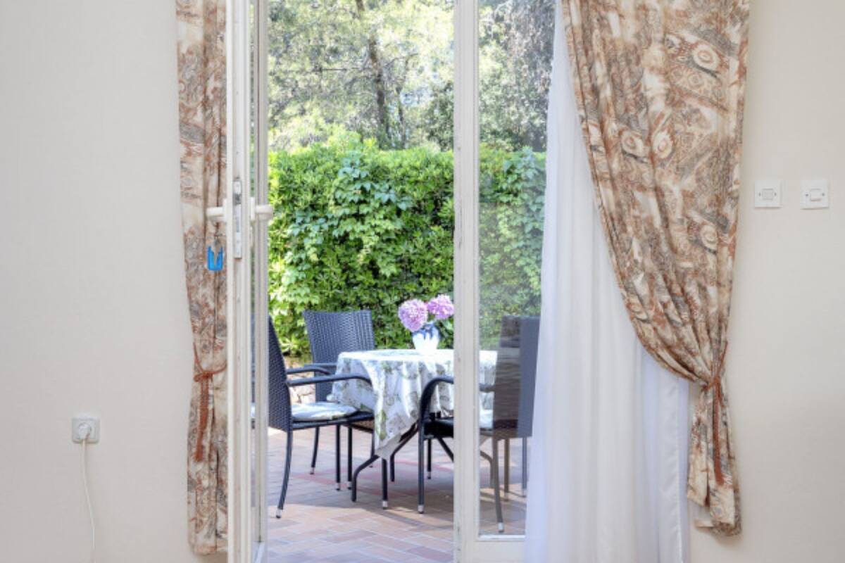 Una stanza con una porta vetrata scorrevole che si apre su un patio esterno con tavolo e sedie, immerso nel verde.