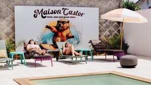 A poolside area with lounge chairs, a small pool, and a large poster advertising 'Maison Castor'. - *Maison Castor* Balneo, pool and billiards! 10 minutes from the ocean! (Mimizan)