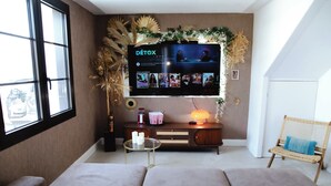 A living room with a large flat-screen TV mounted on the wall, a sofa, a small round table with a lamp, and a wicker chair. - *Maison Castor* Balneo, pool and billiards! 10 minutes from the ocean! (Mimizan)