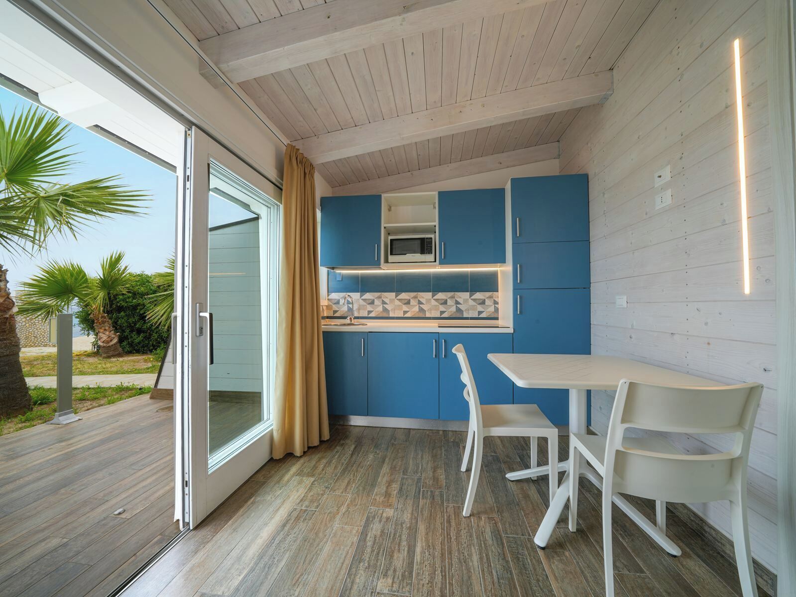 A modern kitchen with blue cabinets, a white countertop, and a microwave. There is a dining area with a white table and chairs.