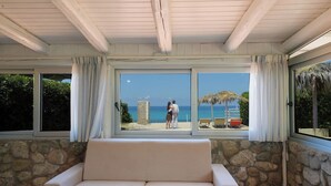 Ein Zimmer mit Steinwand, einem beigefarbenen Sofa und einem großen Fenster mit Blick auf einen Strand, auf dem sich ein Paar und Strandstühle befinden.
