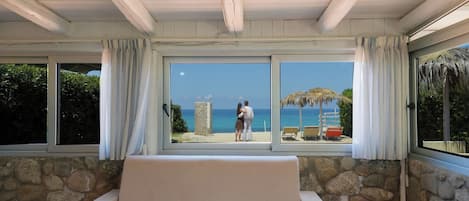 Una habitación con pared de piedra, un sofá color beige y un ventanal con vistas a una playa donde hay una pareja y sillas de playa.