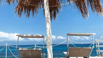 A beach with two wooden lounge chairs under a palm tree, a white fence, and a clear view of the ocean.