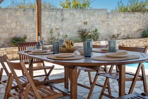 An outdoor dining area with a stone wall backdrop, wooden chairs, a round table, and a bowl of fruit. - Galini villa, private pool , near amenities, Heraklion (Episkopi)