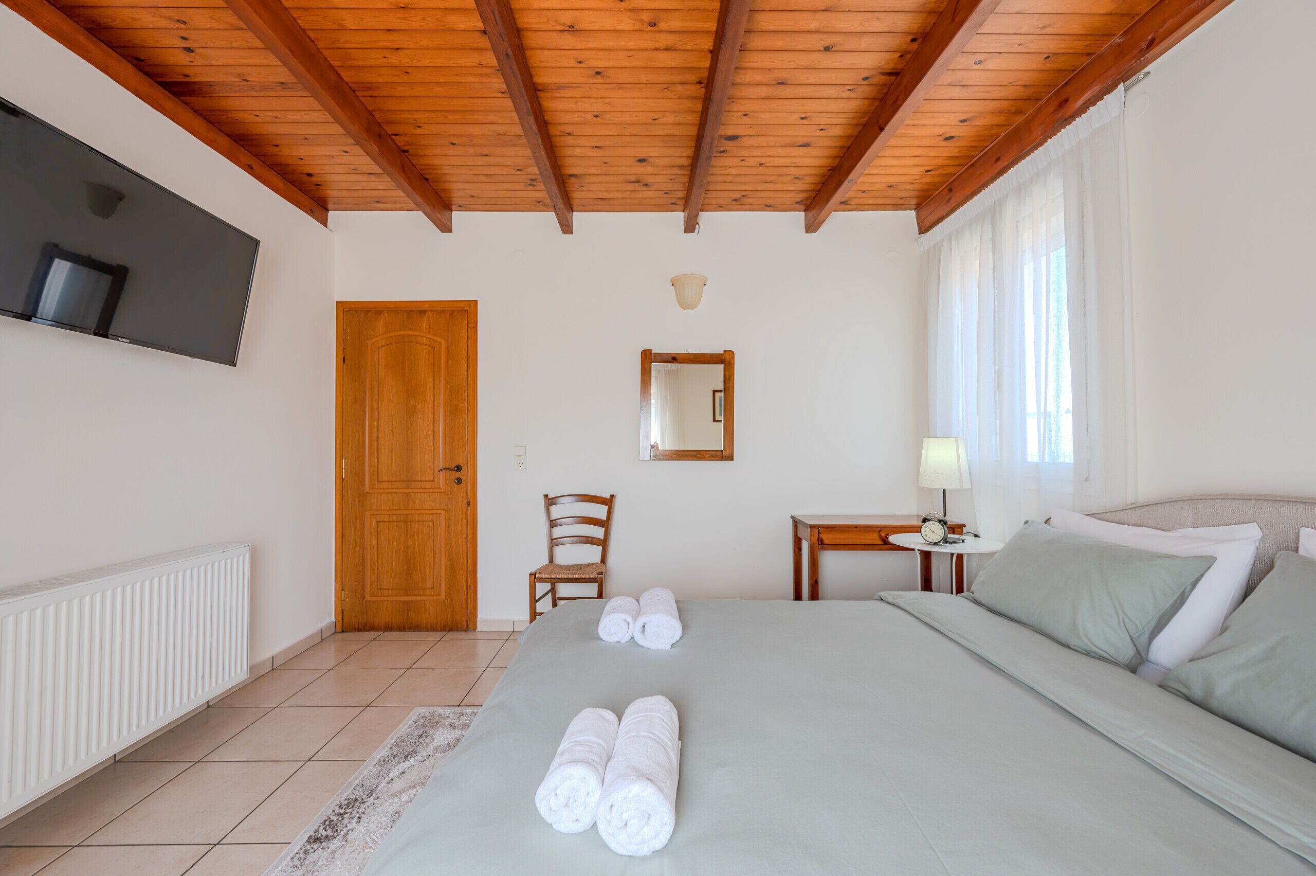 A bedroom with a wooden ceiling, a bed with neatly folded towels, a bedside table with a lamp, a chair, and a TV mounted on the wall.