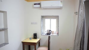 Ein kleines Zimmer mit Bett, Schreibtisch, Stuhl, Fenster mit Aussicht, Klimaanlage und einer Topfpflanze.