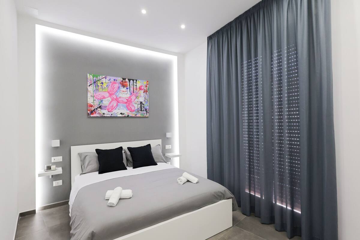 A modern bedroom with a large bed, a colorful abstract painting, and a grey curtain.