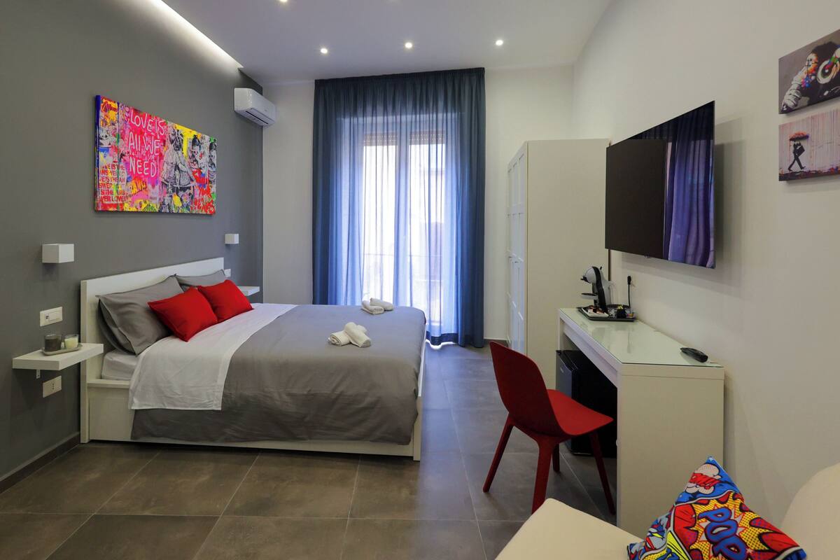 A modern hotel room with a bed, a desk, a TV, and a large window with curtains.