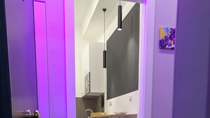 A hallway with purple lighting, a colorful doormat, a kitchen area with a red chair, and a painting on the wall. - Napolitan Pop House (Naples)