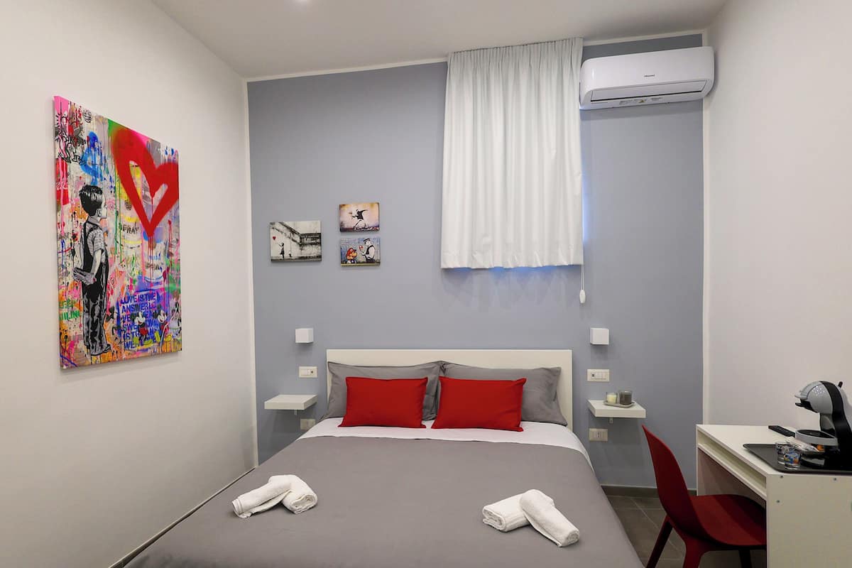 A hotel room with a bed, red pillows, a desk with a chair, and artwork on the wall.