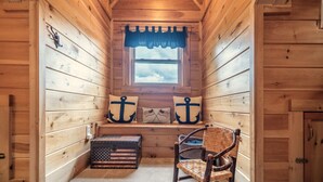 A cabin interior with a wooden interior, a window with a blue valance, a chest of drawers, a wicker chair, and two pillows with anchor designs. - My Happy Place - Lakefront cabin (New Tazewell)