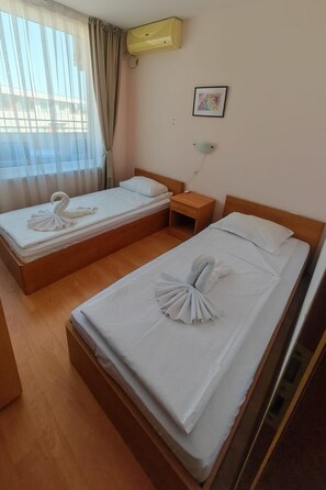 A hotel room with two beds, each with a towel arranged in a swan shape, a bedside table with a lamp, and an air conditioning unit on the wall.