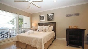A bedroom with a bed, a nightstand, a ceiling fan, a window with blinds, and a wall with two framed pictures and a welcome sign. - 505 Sunsuite (Fripp Island)