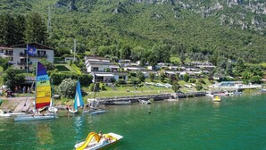 A lakeside view with a sailboat, a speedboat, and a dock with boats. There are buildings in the background and a mountainous landscape. - Directly on the lake with terrace (Tre Capitelli)