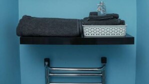 A bathroom with a blue wall, a black shelf holding a folded towel and a white basket, and a chrome towel rack below.