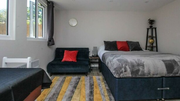 A bedroom with two beds, a bedside table, a chair, a radiator, a clock on the wall, and a window with curtains.