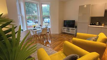 A modern living room with yellow sofas, a dining area with a table and chairs, a TV on a stand, and a kitchen area with cabinets and a countertop.