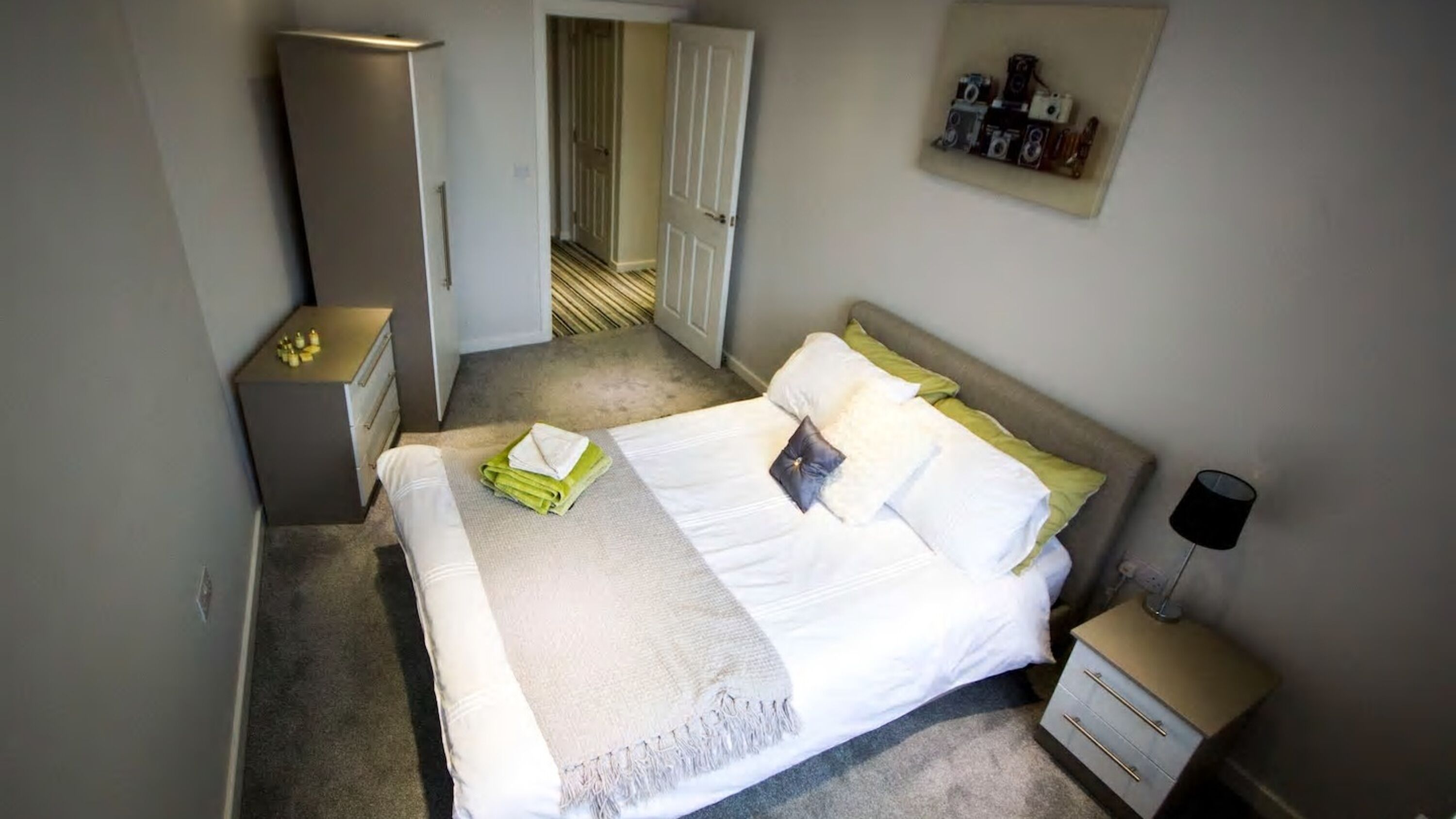 A neatly made bed with a white comforter and pillows, a bedside table with a lamp, and a door leading to another room.