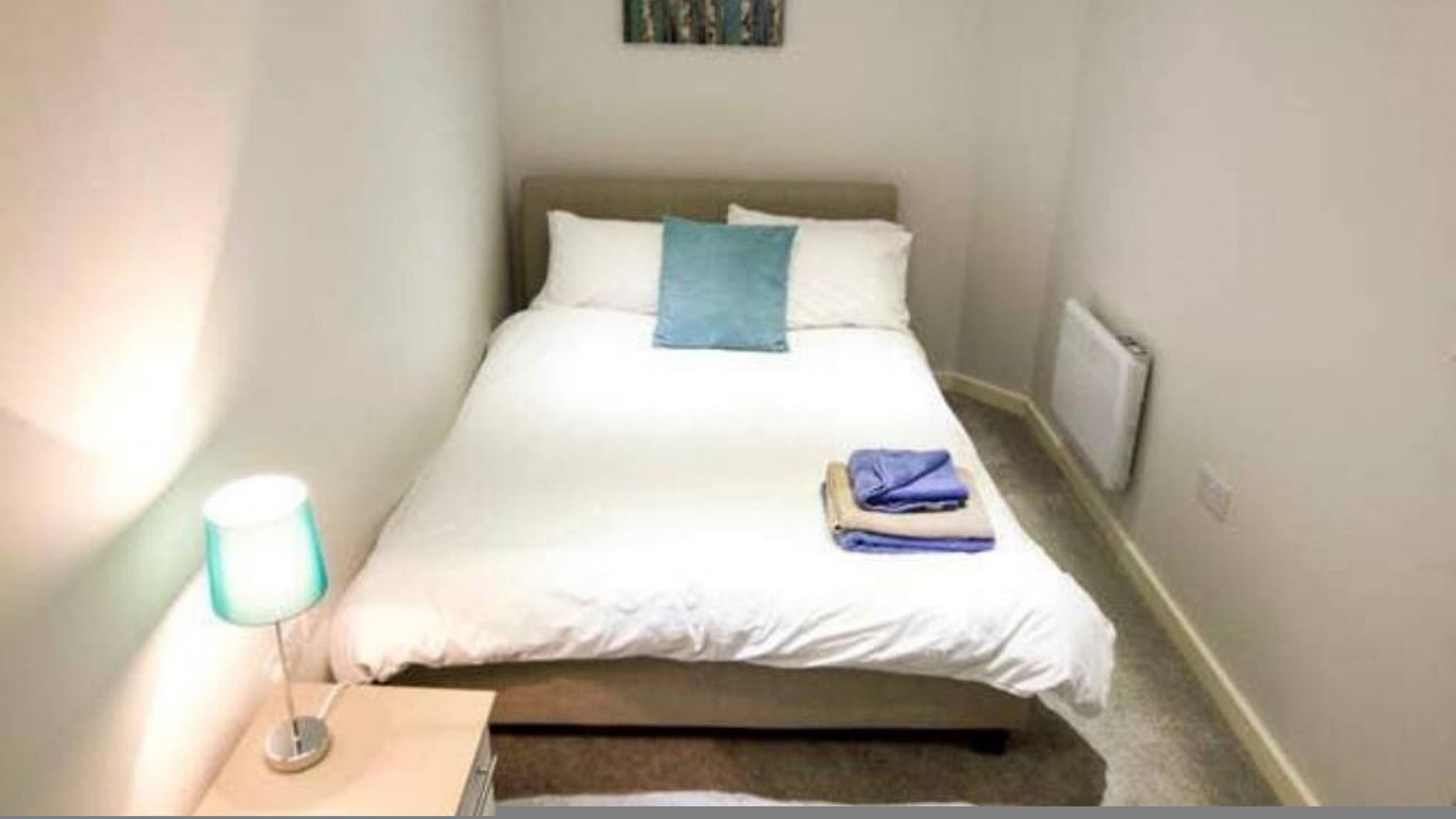 A small, neatly made bed with white bedding and a blue pillow, a bedside lamp, and folded clothes on the floor beside the bed.