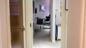 Un pasillo estrecho con una puerta a la izquierda, una mesita con una silla en el centro y una ventana con persianas a la derecha.