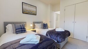 A hotel room with two beds, a nightstand with a lamp, a painting on the wall, and a wardrobe. - Luxury Apartment - Town Centre North Stevenage (Stevenage)