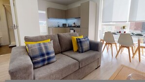 A modern living room with a grey sofa, two yellow and blue pillows, and a dining area with wooden chairs and a table. - Luxury Apartment - Town Centre North Stevenage (Stevenage)