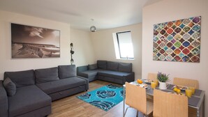 Un soggiorno moderno con un divano angolare grigio, un tavolo da pranzo con sedie, un tappeto floreale blu e un quadro a tema spiaggia sulla parete.