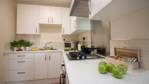 Una cucina moderna con mobili bianchi, piano cottura in acciaio inossidabile e forno a microonde. Sul bancone ci sono mele verdi e un contenitore di legno con utensili da cucina.