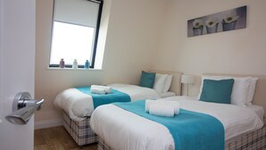 Camera d'albergo con due letti, ognuno con coperta blu e cuscini bianchi. Si vede una finestra con una vista su delle bottiglie sul davanzale e un quadro di fiori appeso al muro.