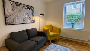 A living room with a black sofa, a yellow armchair, a marble artwork, a floor lamp, and a window with a view of greenery.