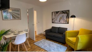 A living room with a grey sofa, a yellow armchair, a dining table set for four, and a painting on the wall.