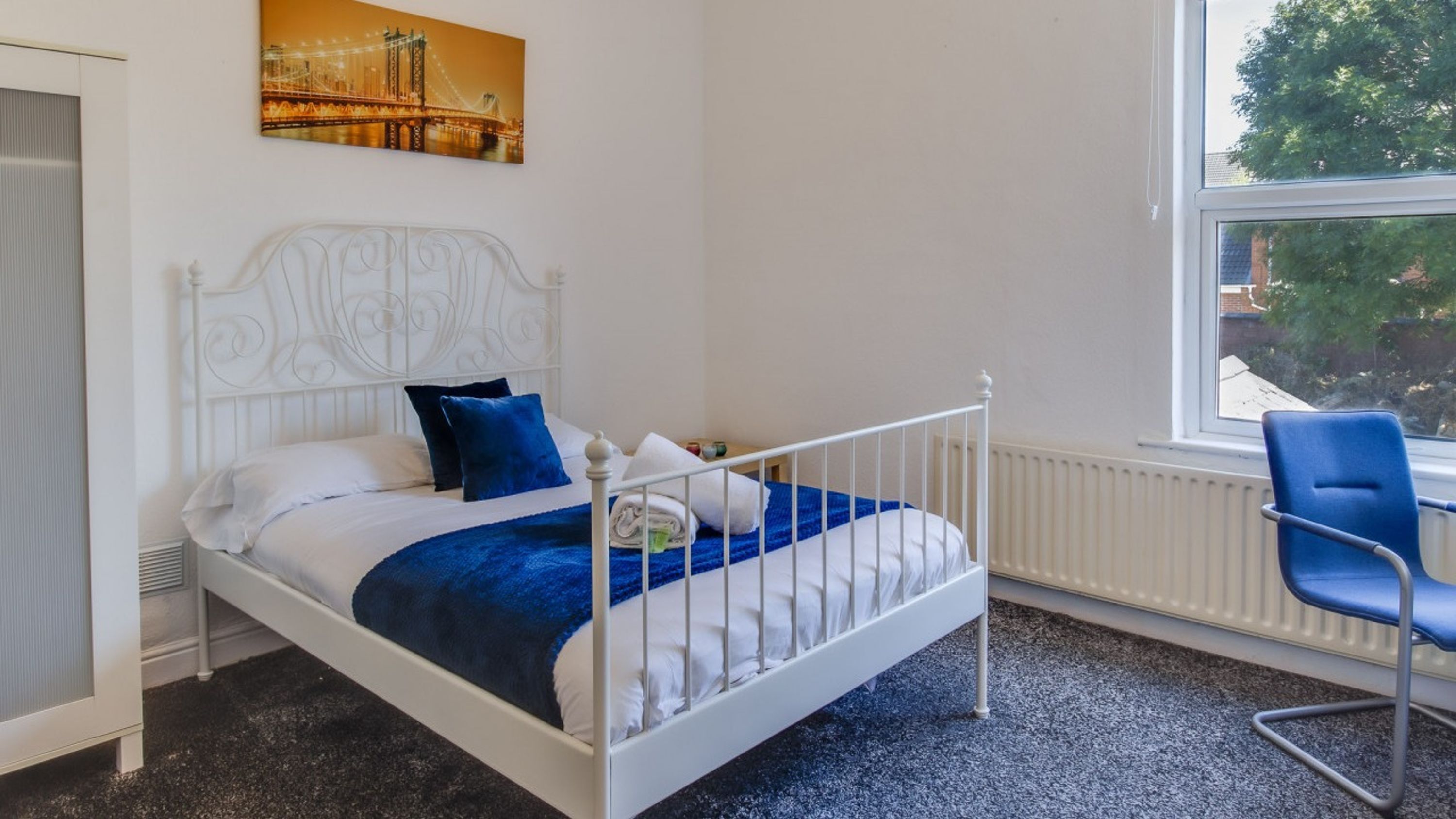 A white metal bed with a blue blanket and pillows, a blue chair, and a window with a view of trees.