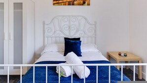 Un lit bien fait, avec des draps blancs et une couverture bleue, une tête de lit blanche et une petite table de chevet en bois sur laquelle se trouvent une tasse et un livre.