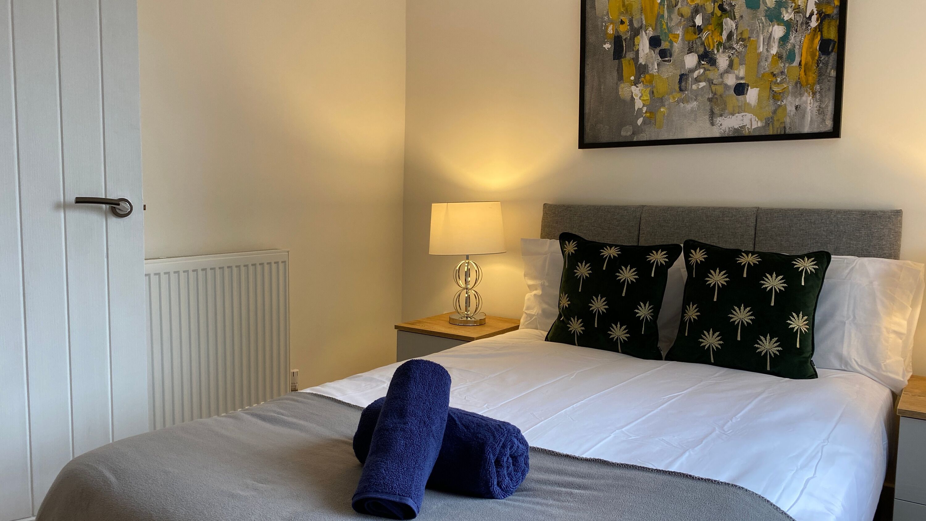A hotel room with a bed, a nightstand with a lamp, a painting on the wall, and a door.