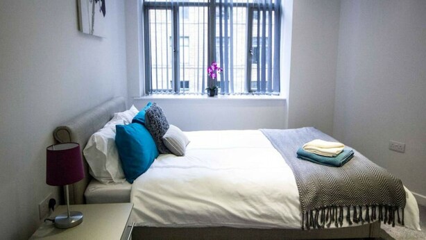 A bedroom with a bed, a bedside table, a lamp, and a window with a view of buildings and a plant.